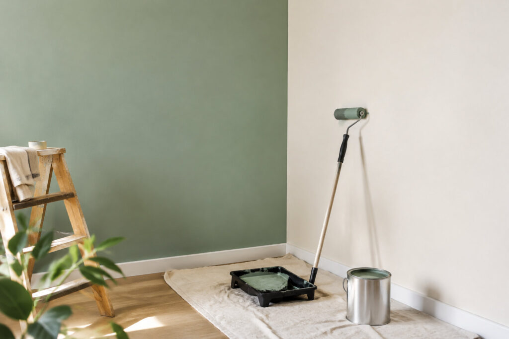 Un rouleau et un bac de peinture verte posés devant un mur bicolore, vert et beige, dans une pièce en cours de rénovation.